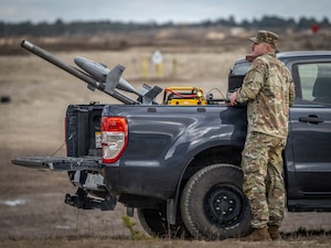 A soldier in a camouflage military uniform stands outdoors near a truck. A device that looks like a missile attached to a pole is mounted in the back of the truck.