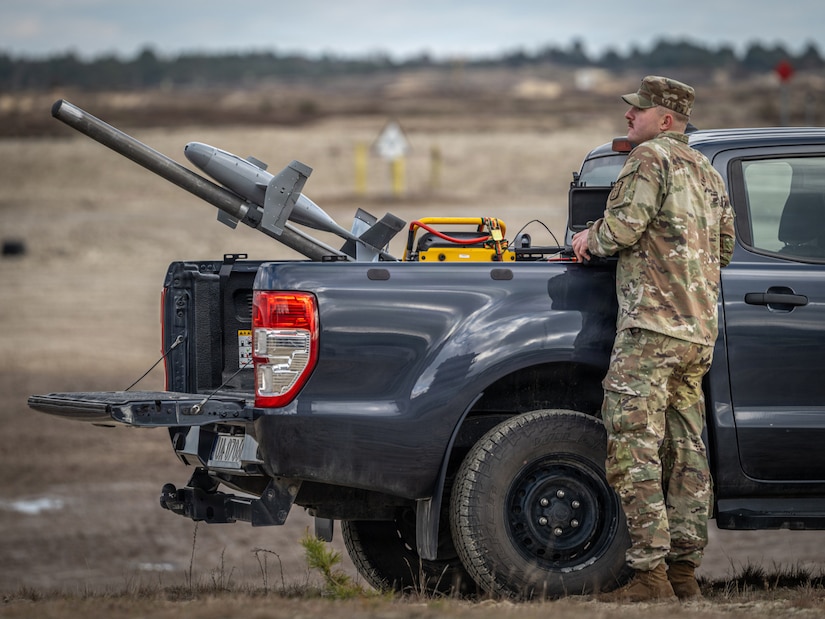 A soldier in a camouflage military uniform stands outdoors near a truck. A device that looks like a missile attached to a pole is mounted in the back of the truck.