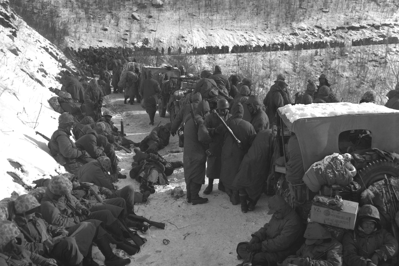 Hundreds of men in cold-weather gear sit and stand alongside a line of military vehicles on a mountainside in the snow.