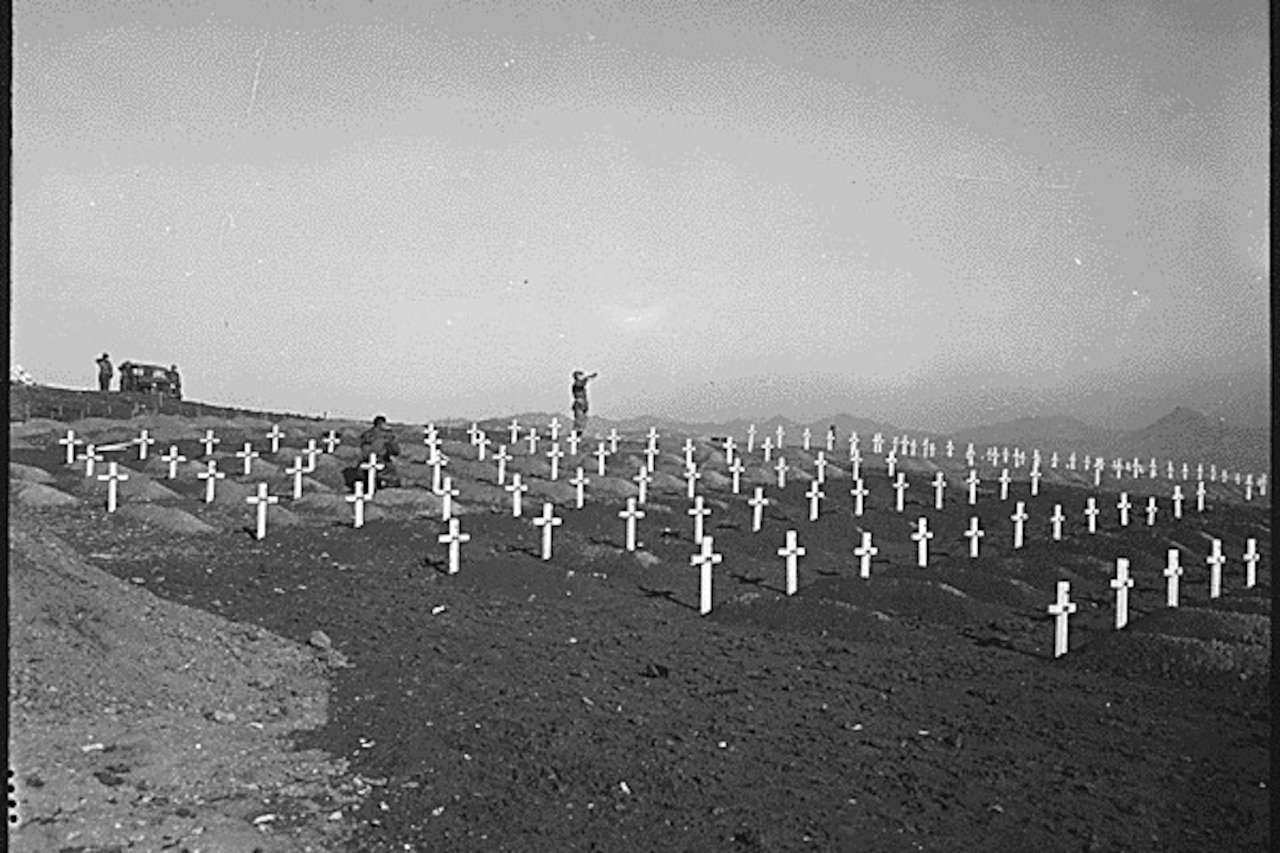 A man plays a bugle in the middle of a graveyard full of white crosses.