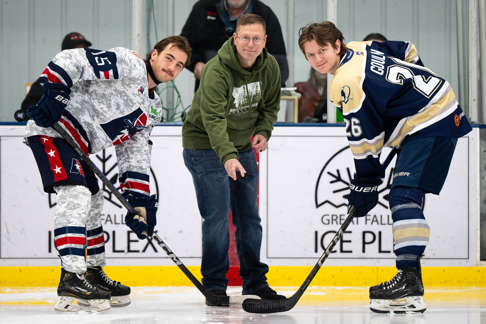 A man in civilian attire holds a hockey puck in between two hockey players.