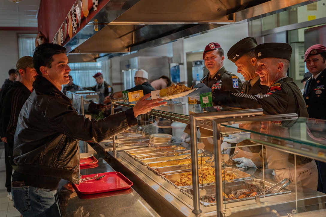 Uniformed soldiers stand behind glass in front of food trays and serve plates full of food to people with red trays.