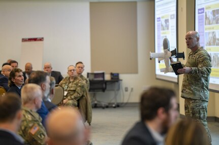 A man in a camouflage military uniform stands in front of a seated audience.