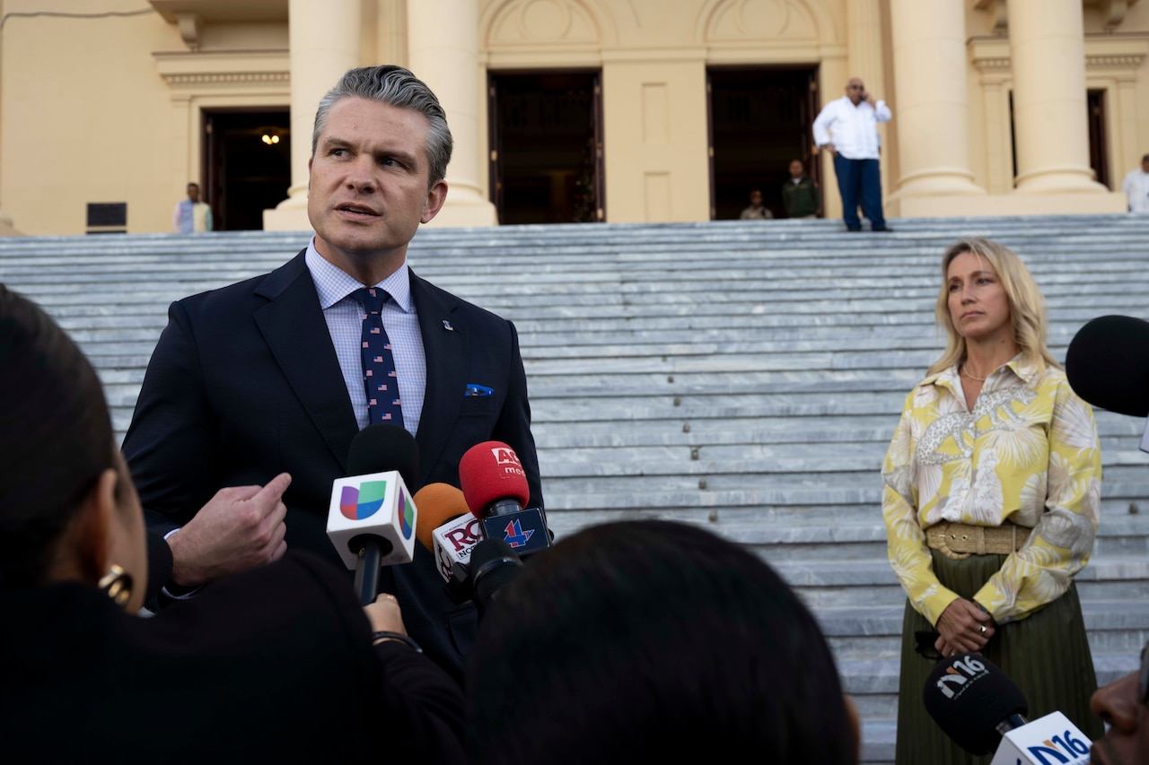 A person wearing a business suit speaks outdoors in front of microphones and other people gathered in the foreground, with another person and a large set of steps in the background.