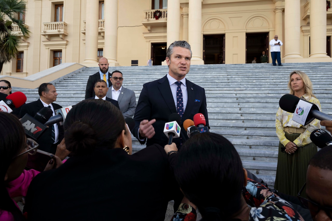 A person wearing a business suit speaks outdoors in front of microphones and other people gathered in the foreground, with more people and a large set of steps in the background.