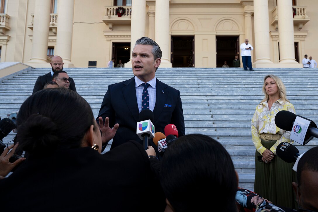 A person wearing a business suit speaks outdoors in front of microphones and other people gathered in the foreground, with more people and a large set of steps in the background.
