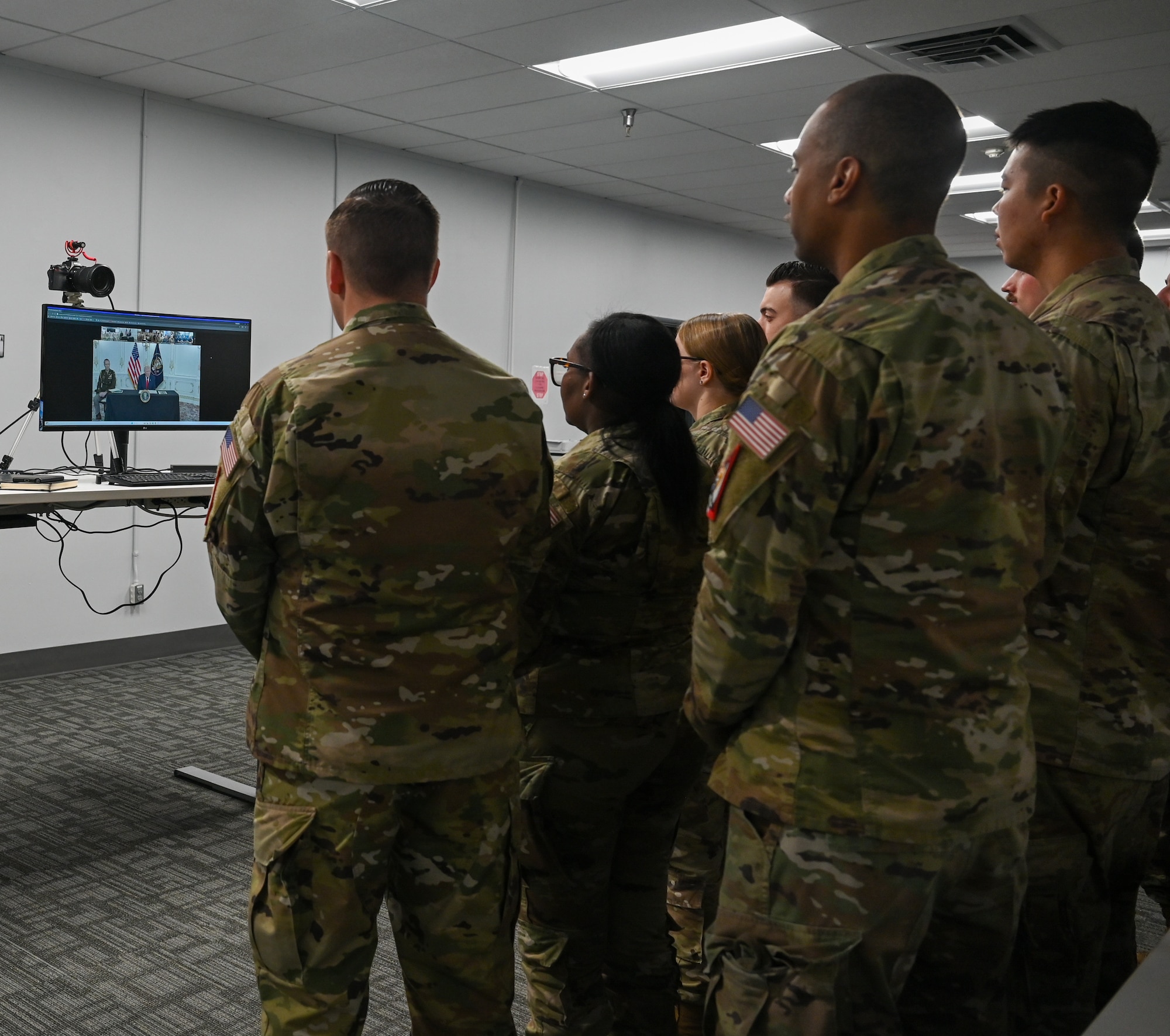 U.S. President Donald J. Trump speaks with service members from across all military branches during a virtual meeting. Service members stand and watch the screen.
