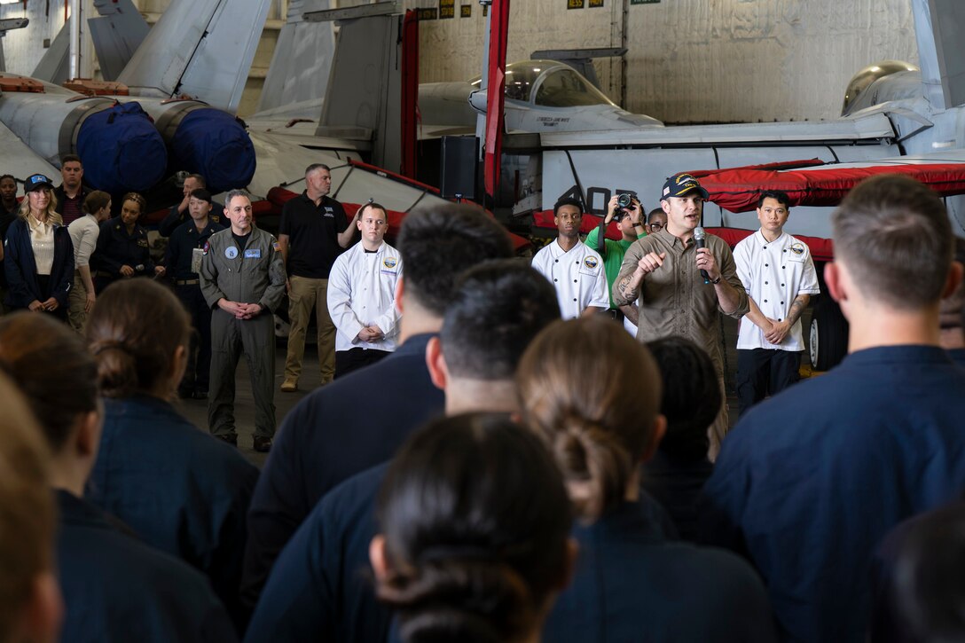 A person in a military flight suit stands in a ship's aircraft hangar holding a microphone while talking to people wearing military coverall uniforms. Many of them have their backs to the camera.