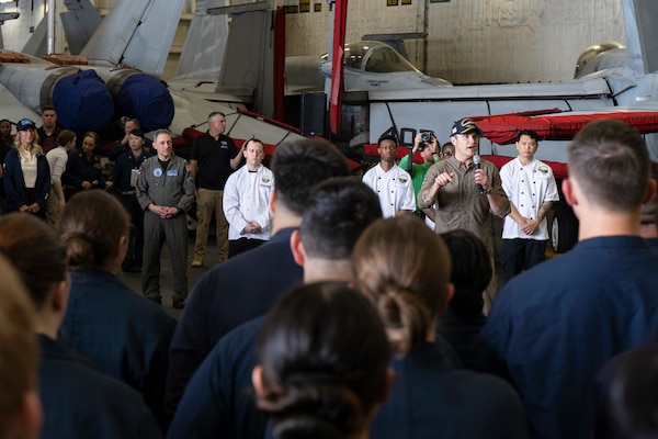 A person in a military flight suit stands in a ship's aircraft hangar holding a microphone while talking to people wearing military coverall uniforms. Many of them have their backs to the camera.