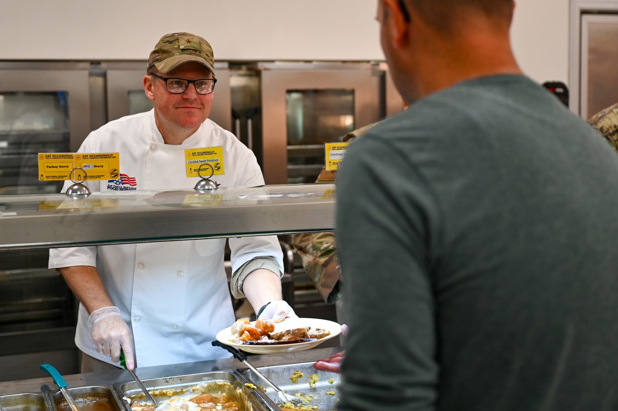 Brig Gen Clayton smiles from behind a serving line as he hands a plate of food to someone in the foreground