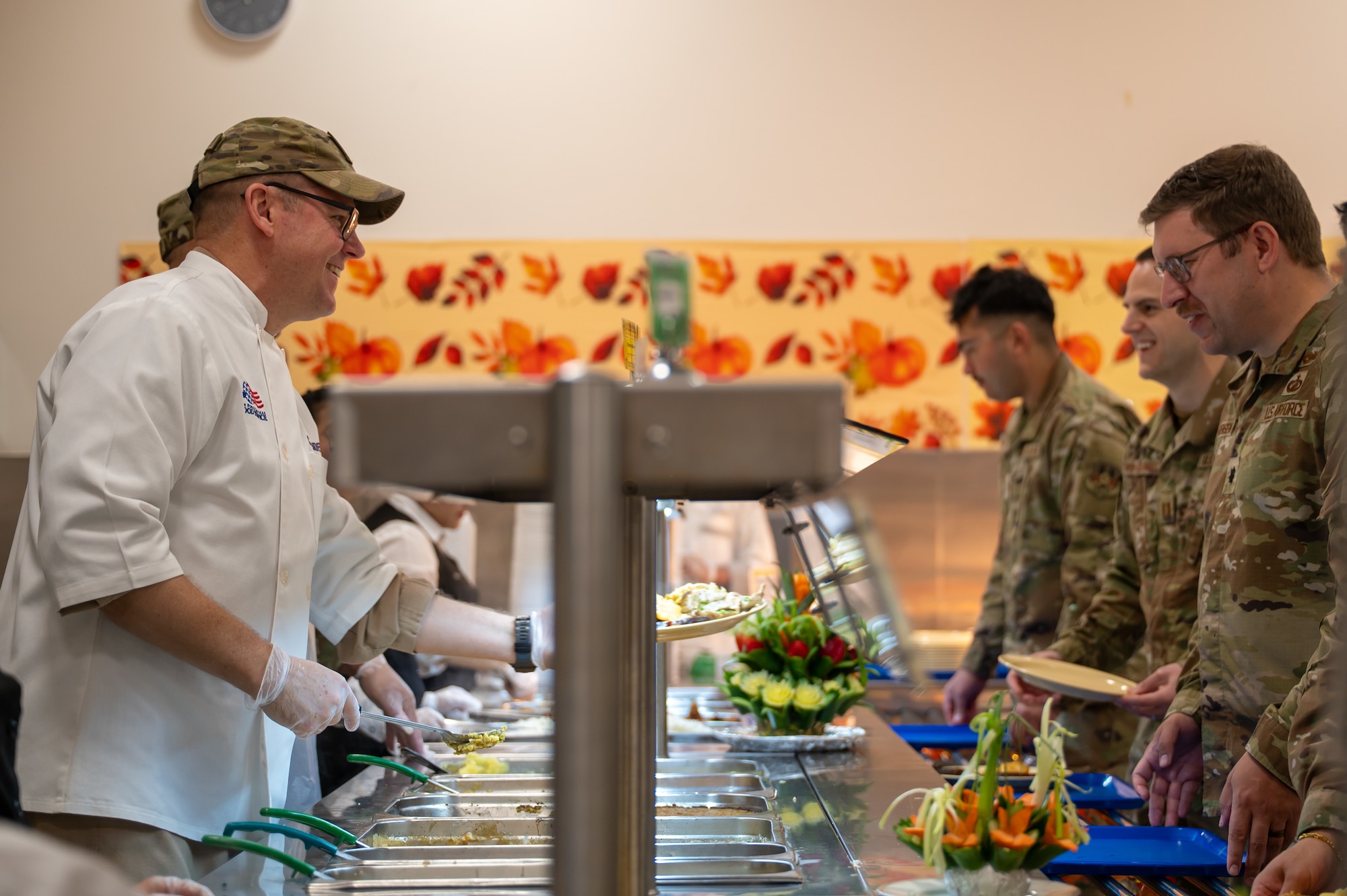Service members stand on either side of a serving line. Brig Gen Clayton, on the left side, smiles as he hands a plate to someone on the right side.
