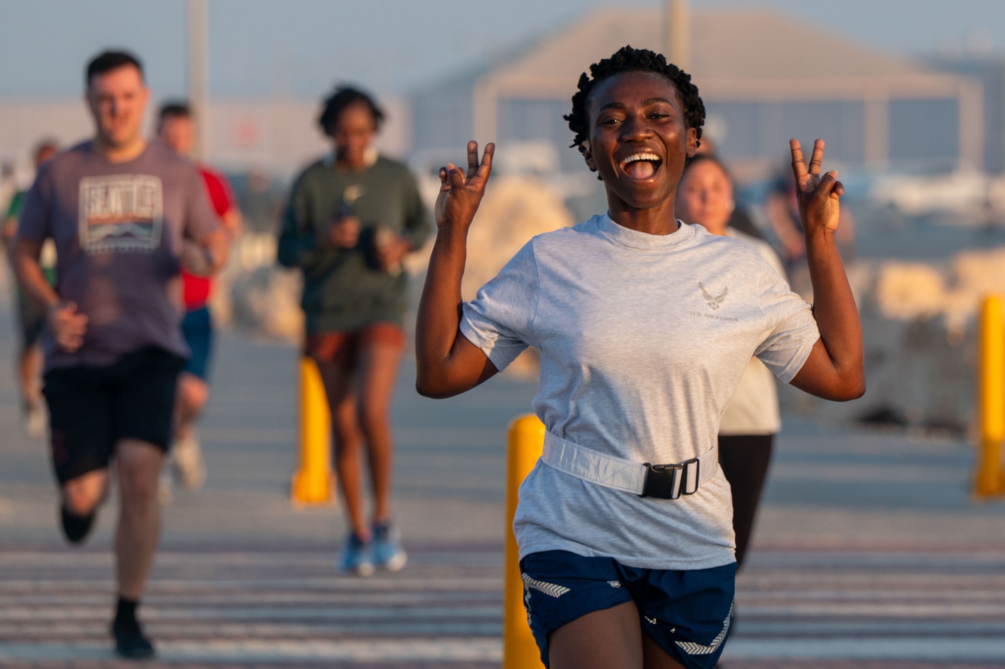 A woman in Air Force physical training gear smiles and makes two peace signs with her hands while running. Other runners are in the background out of focus.
