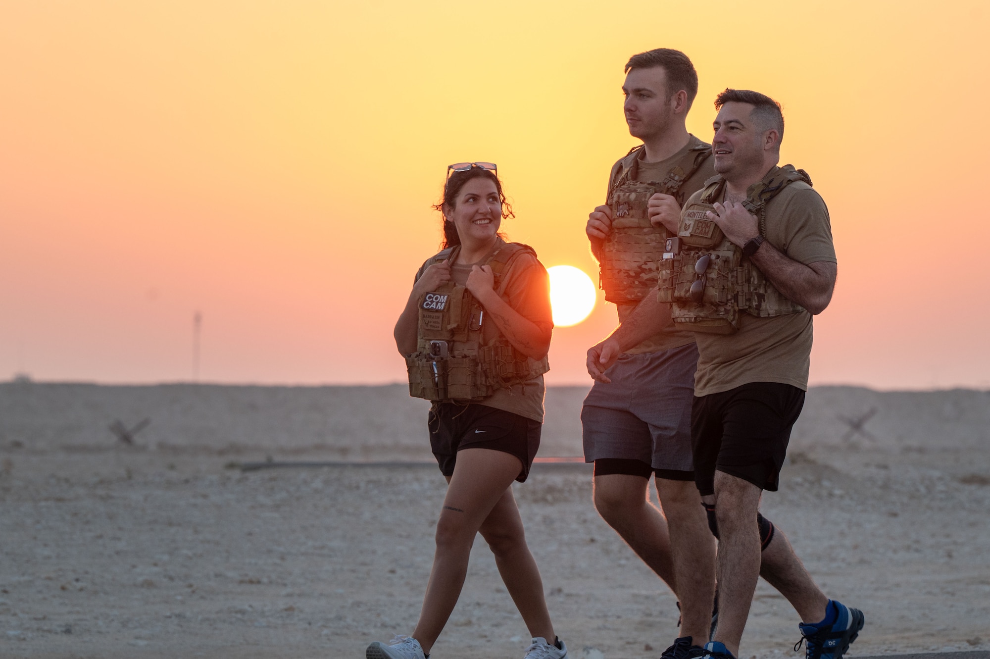 Three people wearing tactical vests walk along a road. The sun, low and orange, illumintates behind them