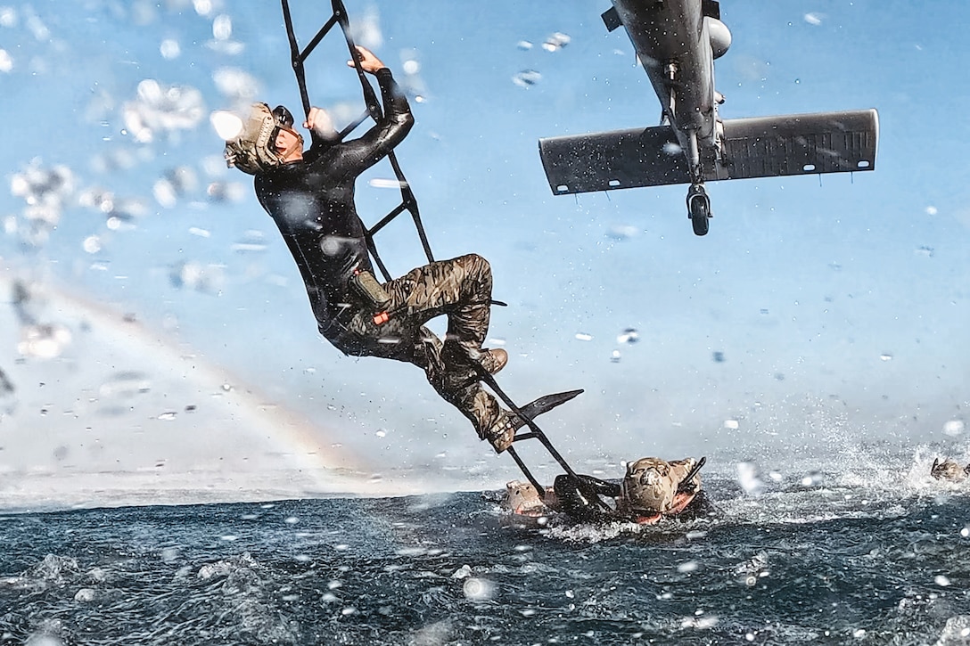 Water splashes in the foreground as a sailor climbs a ladder out of a body of water into a helicopter while a rainbow appears on the left.