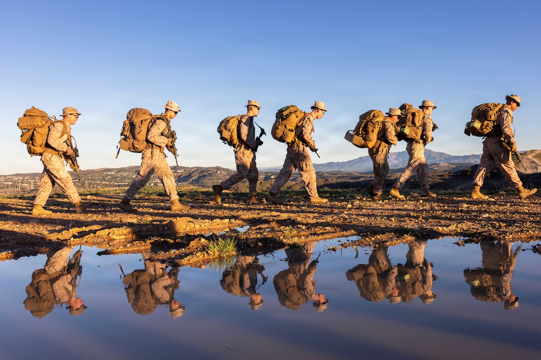 Marines carrying backpacks hike in formation near mountains as their reflection is seen in a puddle of water in the foreground.