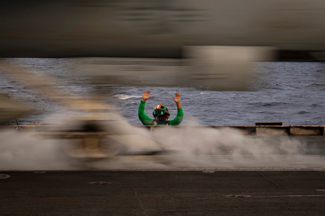 A sailor raises their hands as a blurred aircraft swiftly launches from a ship leaving behind clouds of smoke.