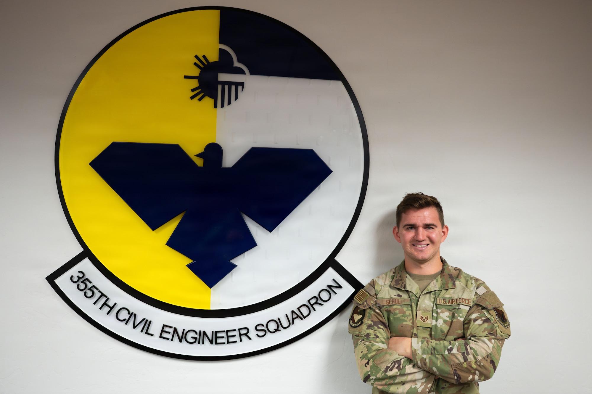 U.S. Air Force Staff Sgt. Joshua Schul, 355th Civil Engineer Squadron unit deployment manager, poses by the 355th CES crest at Davis-Monthan Air Force Base, Arizona, Sept. 26, 2025. The 355th CES is responsible for many of the behind-the scenes base functions including maintenance of all facilities, properly functioning utilities, unaccompanied housing and environmental management. (U.S. Air Force photo by Senior Airman Jasmyne Bridgers-Matos)