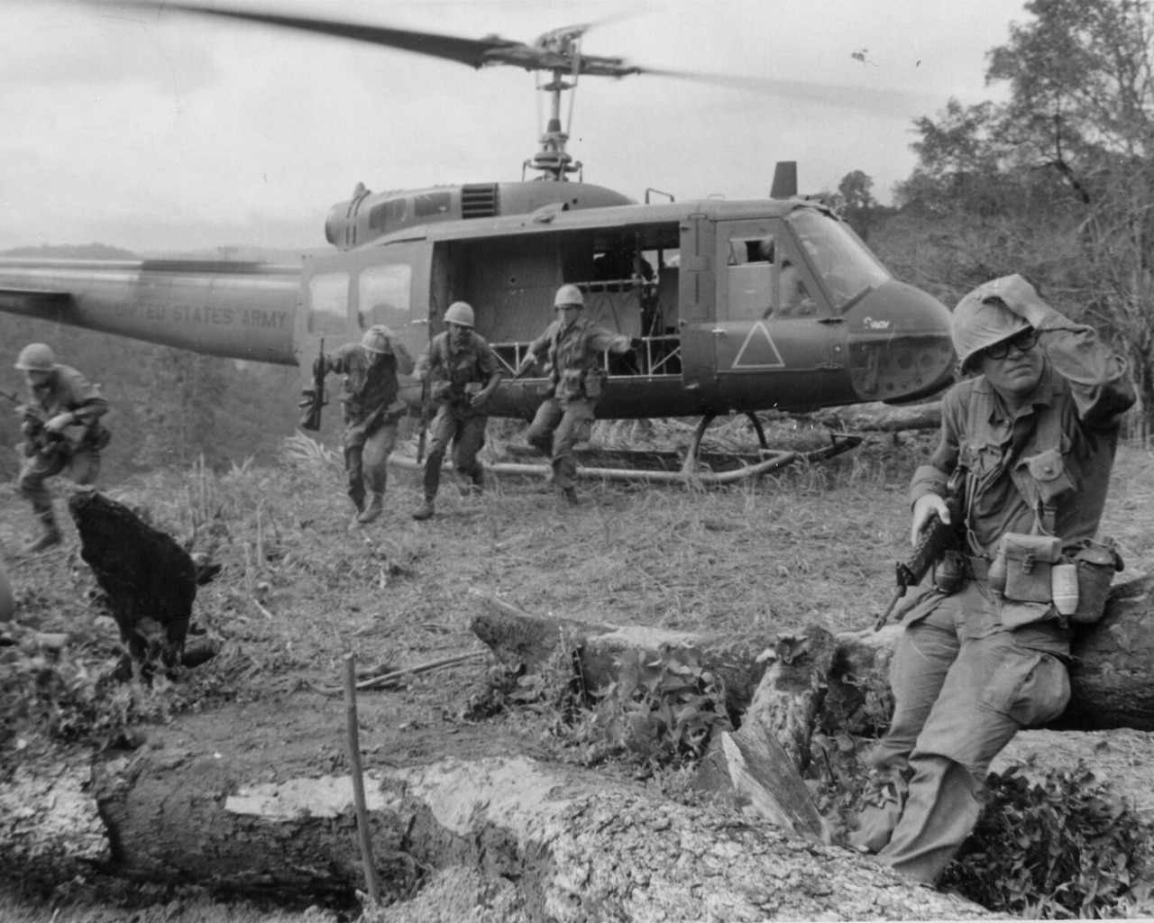 Four men wearing military combat gear move away from a helicopter hovering just off the ground. In the forefront, another man in similar attire sits on a log.