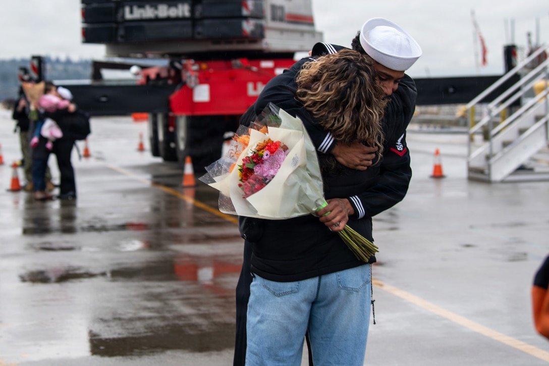 A sailor hugs a person in civilian attire at a wet paved area on a cloudy day, with other people and large construction equipment in the background.