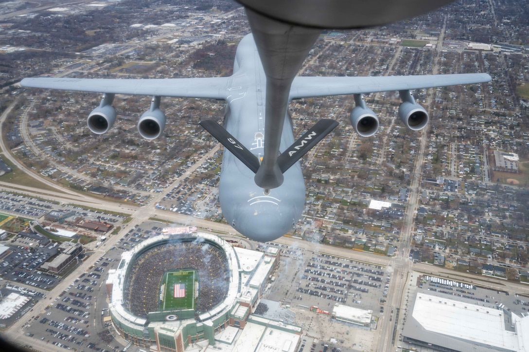 A large airplane trailing another aircraft flies over an urban area and stadium with an American flag on a green field during daytime.
