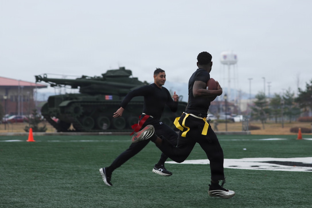 A soldier wearing workout attire and carrying a football in one arm runs away from another soldier in similar attire on a green field during daytime, with two orange cones, a tank and a water tower in the background.
