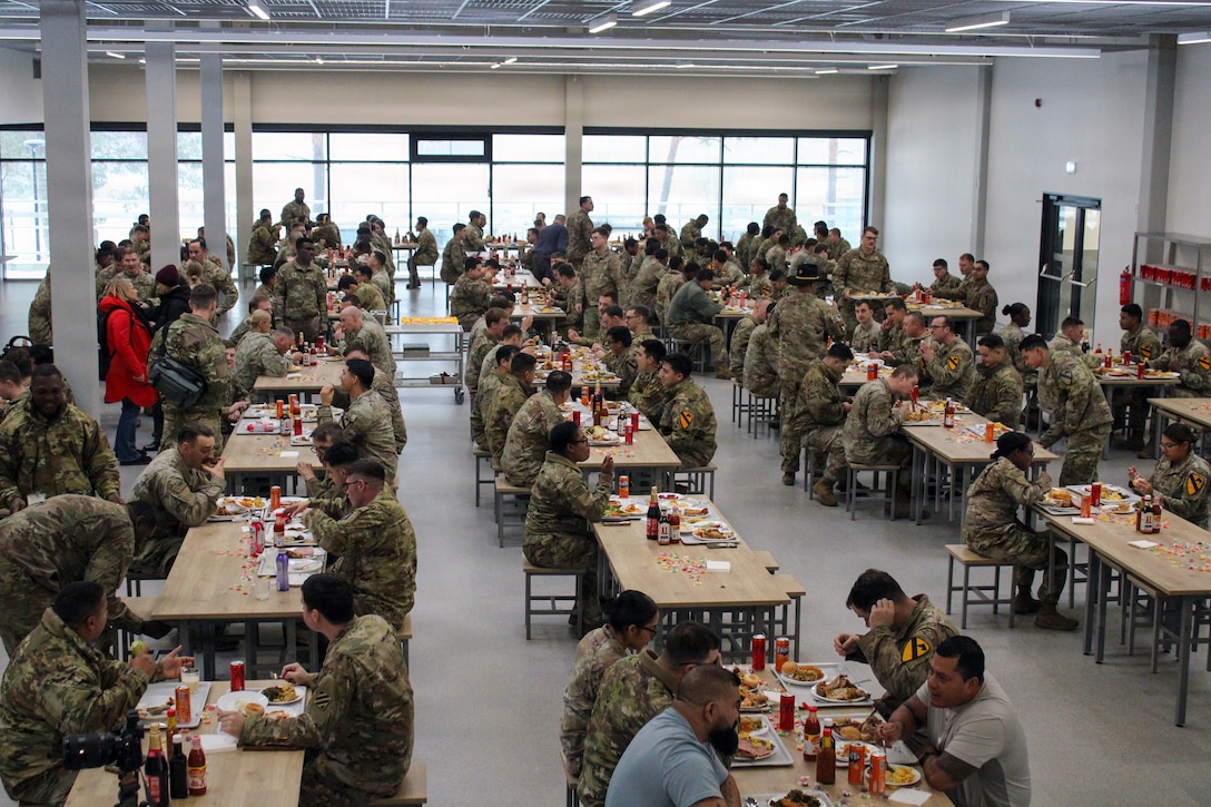 Soldiers sit together at long tables in a dining facility sharing a meal on metal trays.