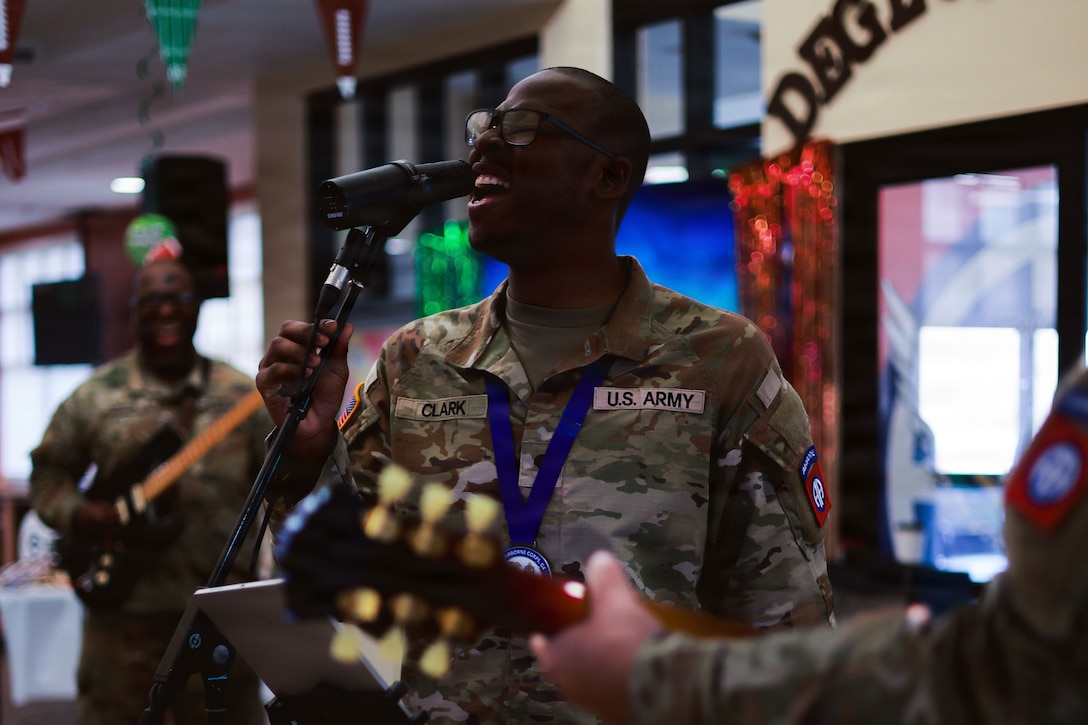 A soldier sings into a microphone beside other band members playing guitars during an indoor event.