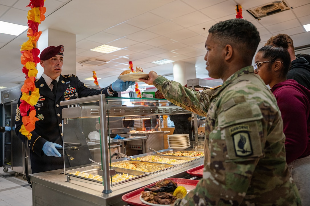 Soldiers sit together at long tables in a dining facility sharing a meal on metal trays.