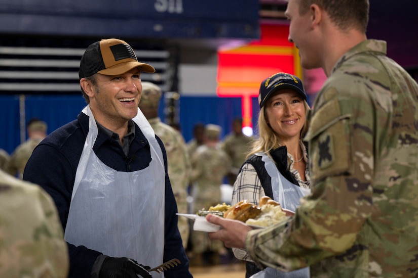 Two people in civilian clothing and plastic aprons smile while serving food to a person in a military camouflage uniform holding a plate of food.