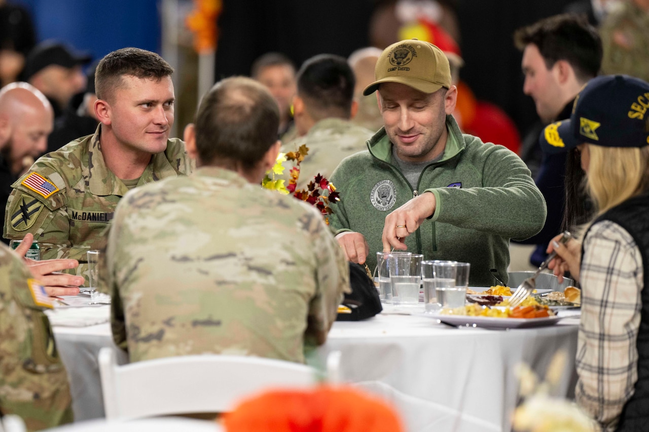 A person in civilian clothes and a hat cuts food while seated at a table with another person in civilian clothes and several individuals in military camouflage uniforms. The people are all eating food at the table.