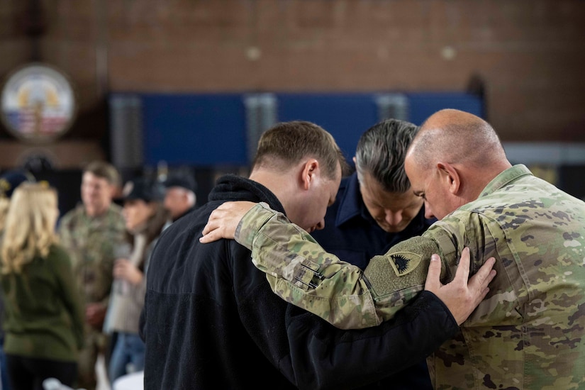 Two people in civilian clothes and one person in a camouflage uniform stand in a circle with their heads bowed and hands on one another's shoulders.