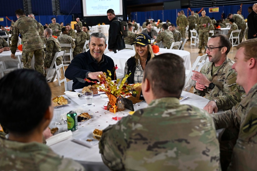 People in civilian clothes sit around a table with several people in military camouflage uniforms as they share a meal together.