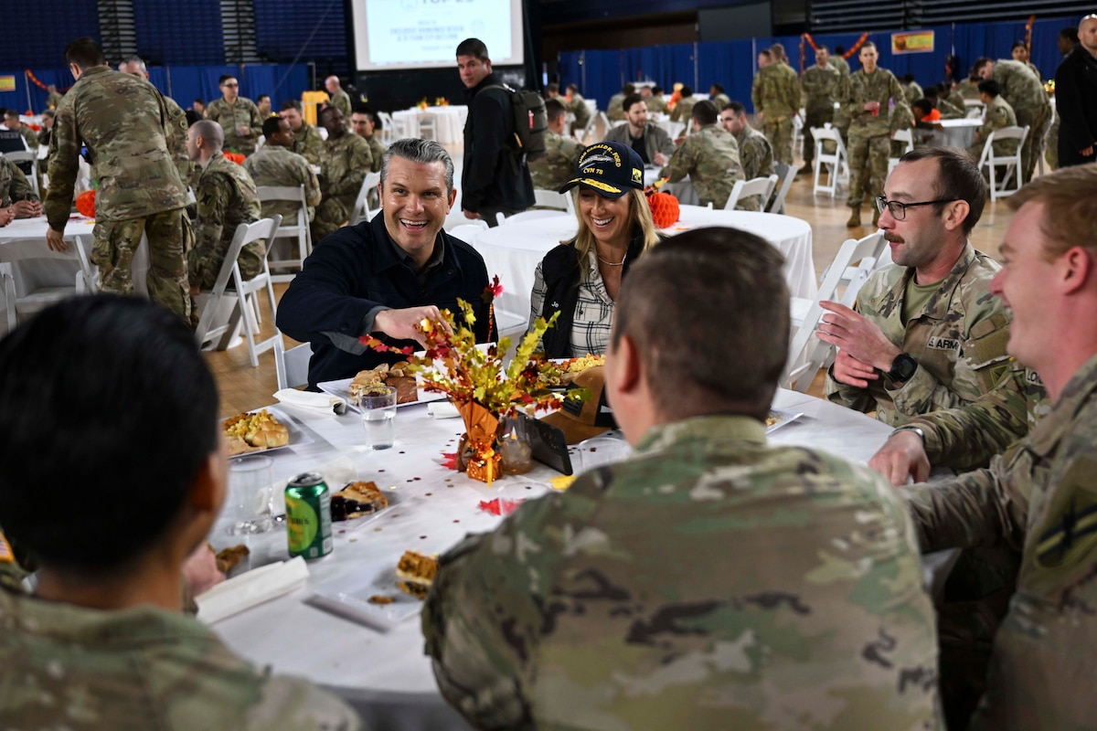 People in civilian clothes sit around a table with several people in military camouflage uniforms as they share a meal together.