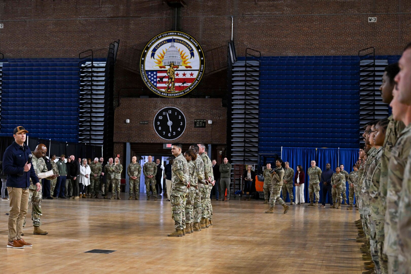 A person in civilian clothing stands in front of a formation of uniformed military personnel in a large arena.