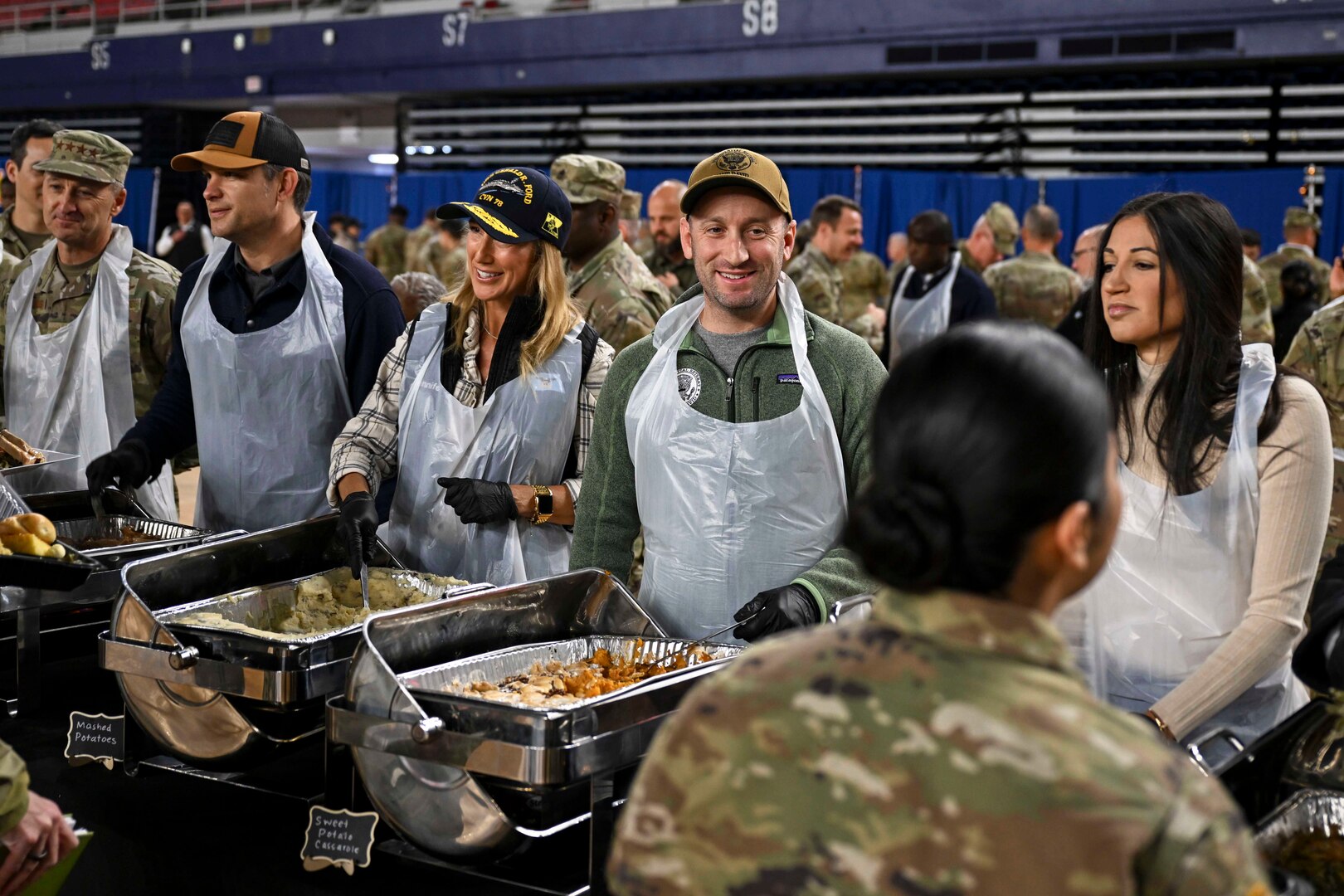 Several individuals in civilian clothing and plastic aprons serve food to uniformed service members.