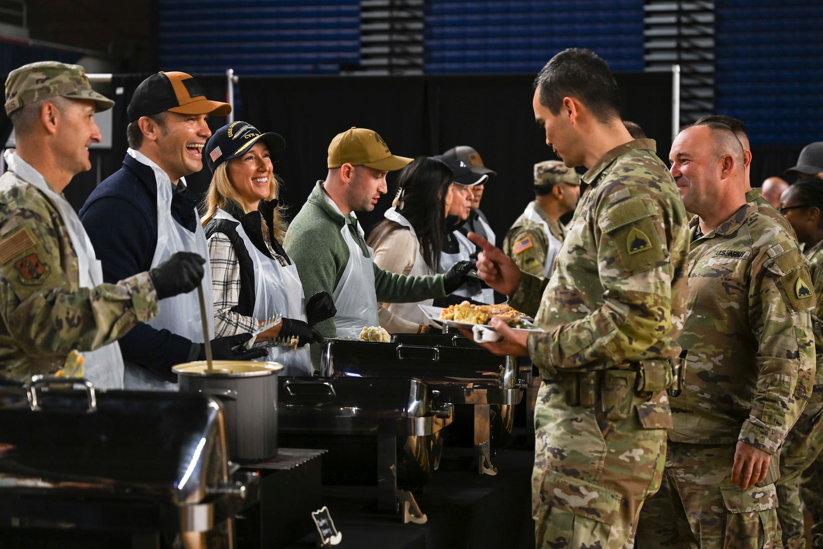 Several individuals in civilian clothing and one in a military camouflage uniform serve food to uniformed service members.