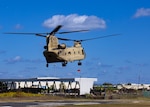 A Chinook helicopter hovers above the ground with blue skis behind it.