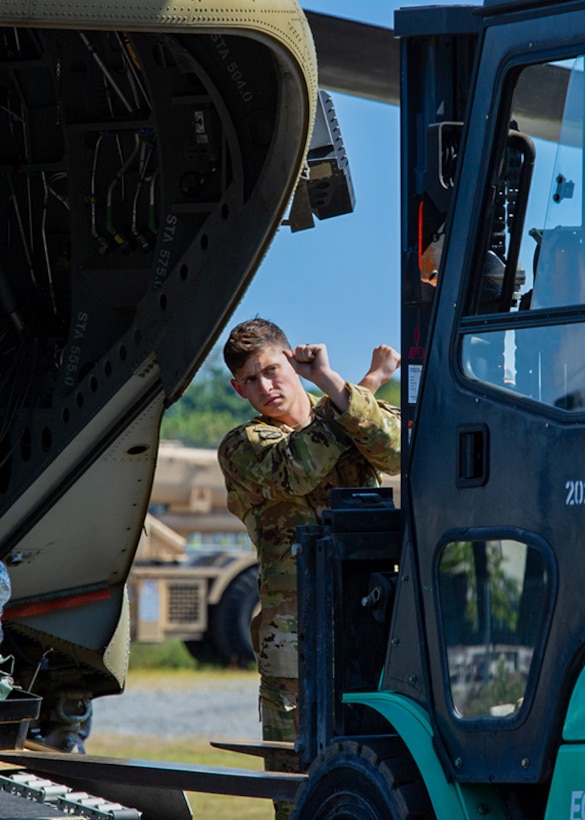 A Soldier in a camo uniform holds up his arms and crosses them in front of himself as a signal to a forklift operator.