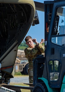 A Soldier in a camo uniform holds up his arms and crosses them in front of himself as a signal to a forklift operator.