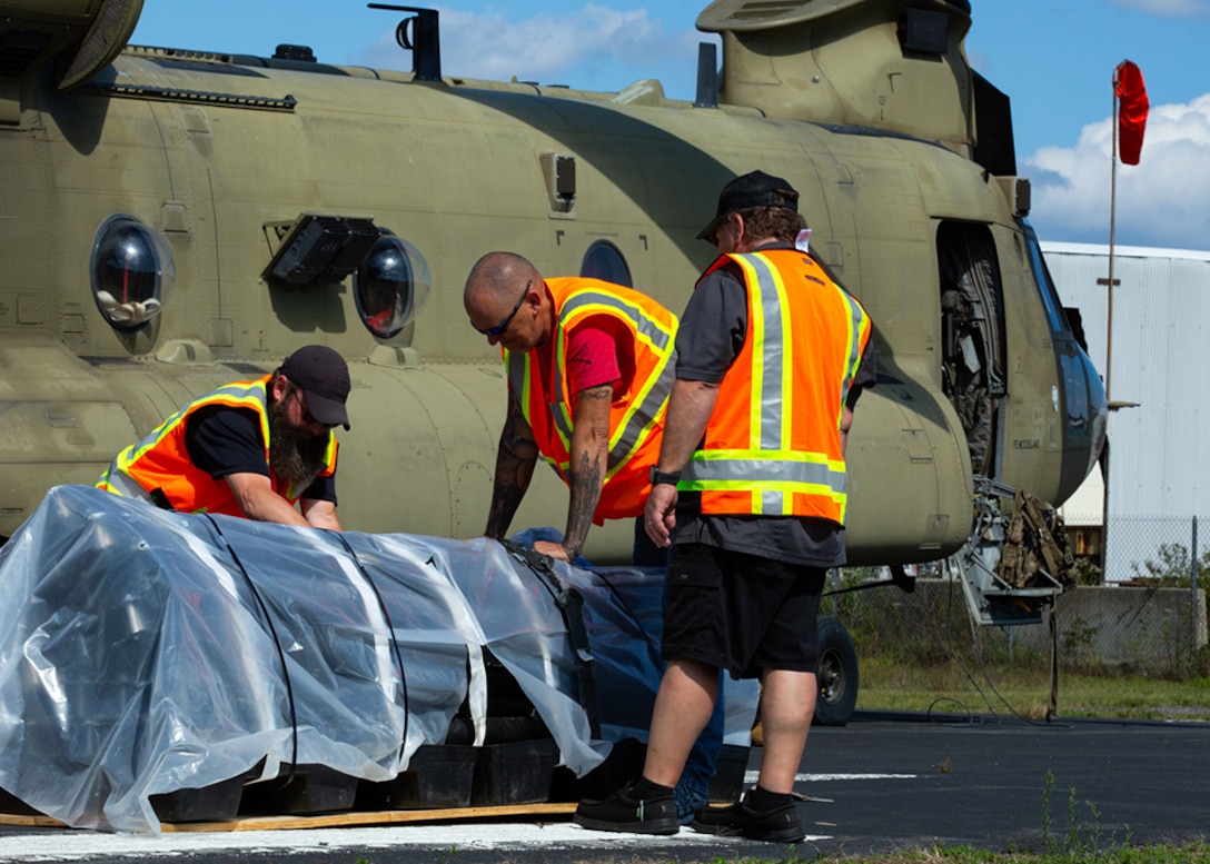 Three DLA Civilians inspect excess property sitting outside on the ground and covered in plastic. a cargo helicopter sits in the background.