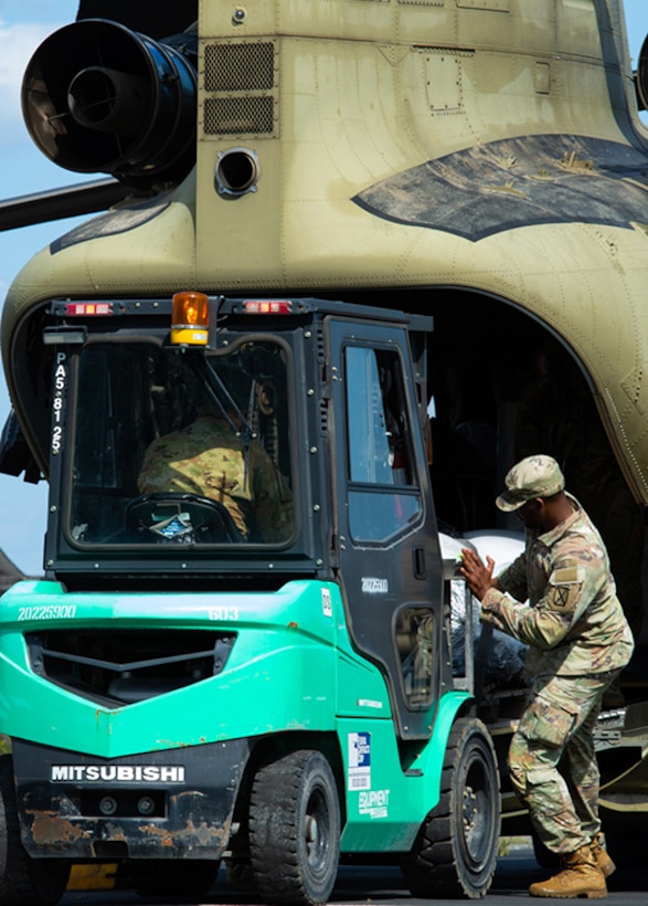 A bright green forklift sits at the back of a Chinook being loaded with cargo from the helicopter by a Soldier in camo uniform.
