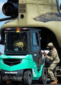 A bright green forklift sits at the back of a Chinook being loaded with cargo from the helicopter by a Soldier in camo uniform.