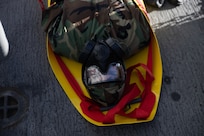 A United States Soldier in full camouflage and gas mask lays on a yellow gurney outside at a training event.