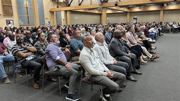 A group of people are seated in an auditorium to listen to a live stream of a town hall broadcast.