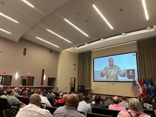 People sitting in an auditorium watching a person speak on a large screen.