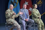 Two men and one woman sit on a stage with military flags behind them.