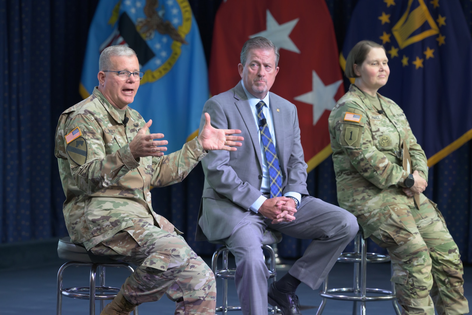 Two men and one woman sit on a stage with military flags behind them.