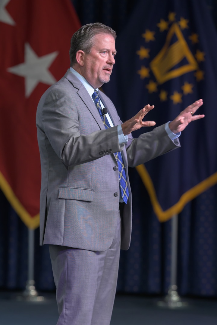 A man is standing on a stage with military flags behind him.