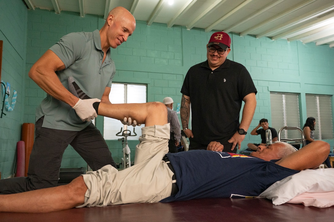 U.S. Army Reserve Maj. Collier Amundson, left, Army Forces Medical Detachment Chief of Rehabilitation, moves a La Paz Honduran local, Rito Chevez’ leg, while Spc. Gabriel Rabell, Army Forces Battalion Military Police, translates at Centro de Rehabilitacion Integral de Comayagua, Comayagua, Honduras, Sept. 17, 2025. Chevez almost lost his leg after he experienced a work accident with a machete. After seeing Amundson for three months, Chevez has regained his ability to walk after his accident. Joint Task Force-Bravo medical providers, like Amundson, routinely conduct local medical missions, providing an opportunity for service members to engage with the local community and improve regional stability. (U.S. Air Force photo by Staff Sgt. Sadie Colbert)