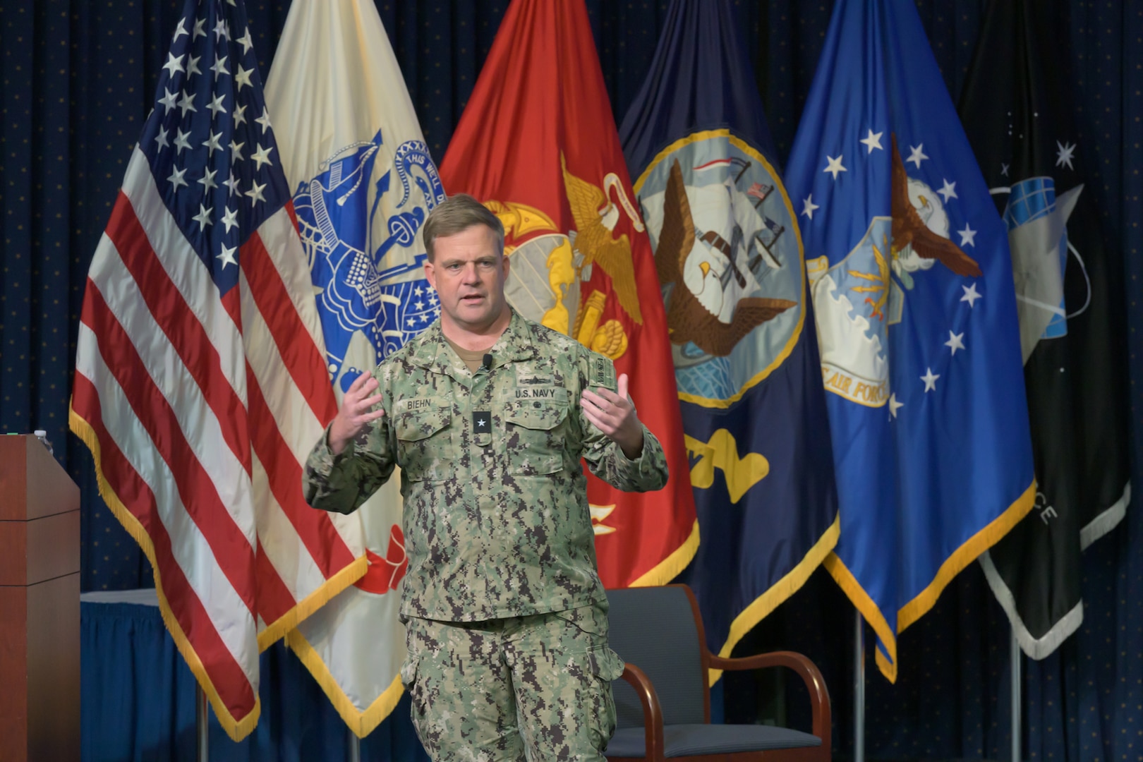 A Navy rear admiral talks on a stage in an auditorium.